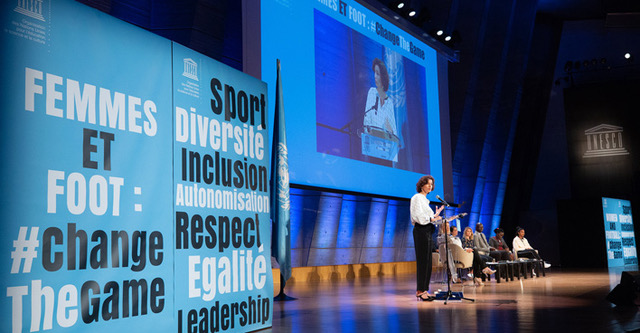 Man sieht eine Veranstaltung der UNESCO, zu sehen ist eine Bühne mit einer Frau am Mikrofon und um HIntergrund sitzen Menschen auf Stühlen. Links sieht man ein Plakat: Femmes et foot: #changeTheGame Sport Diversité Inclusion Autonomisation Respect Egalité Leadership